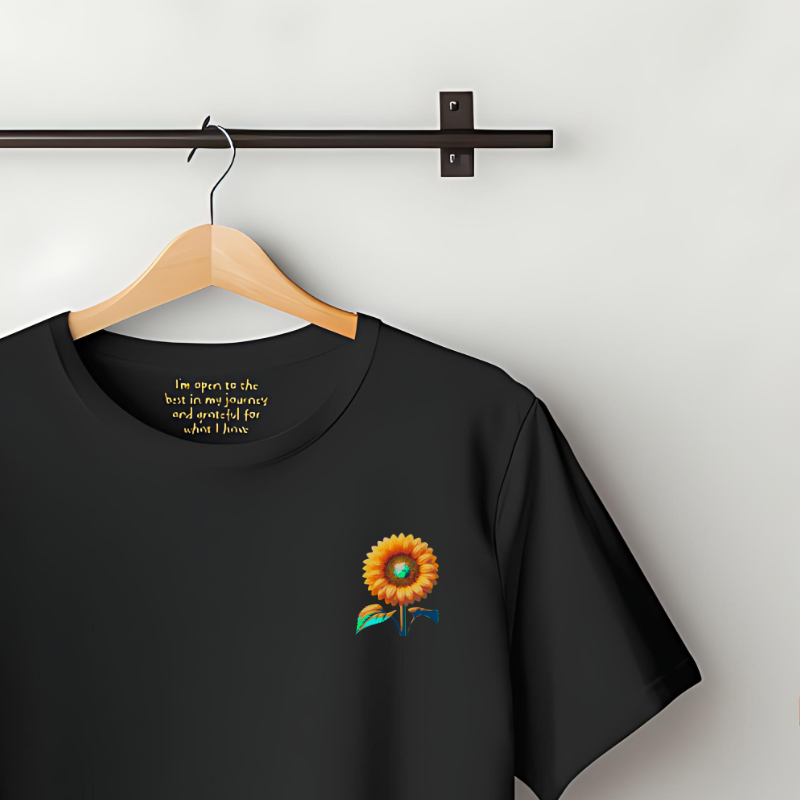 100% Organic Cotton Black t-shirt featuring a bold sunflower graphic on the chest. Text inside the collar reads ""i'm open to the best in my journey and grateful for what I have"